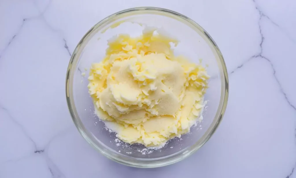 Mélange de beurre crémeux et de sucre pour frangipane, présentant une texture jaune pâle et moelleuse dans un bol en verre transparent sur une surface en marbre