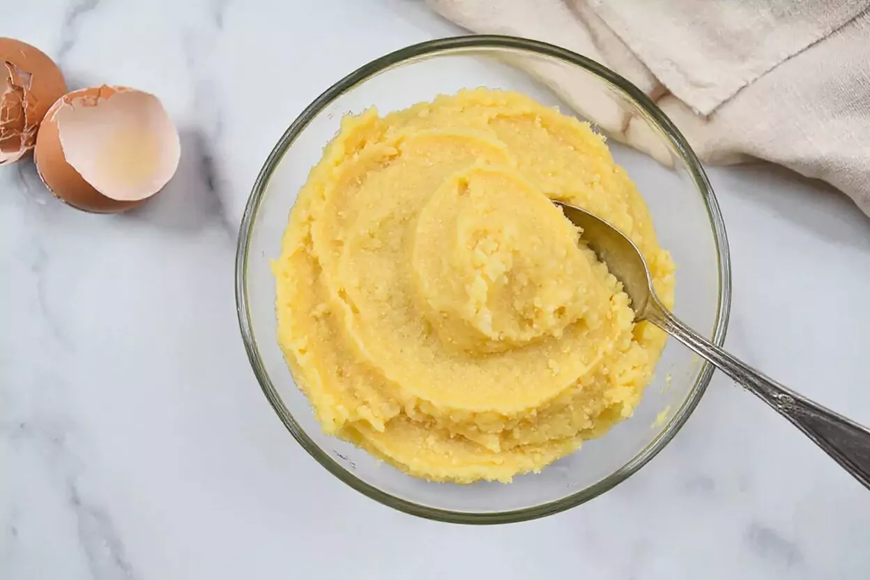 Crème d'amandes frangipane finie dans un bol en verre, montrant une garniture de pâte jaune pâle lisse et cohésive avec des amandes éparpillées et des morceaux de coquille d'œuf sur une surface en marbre